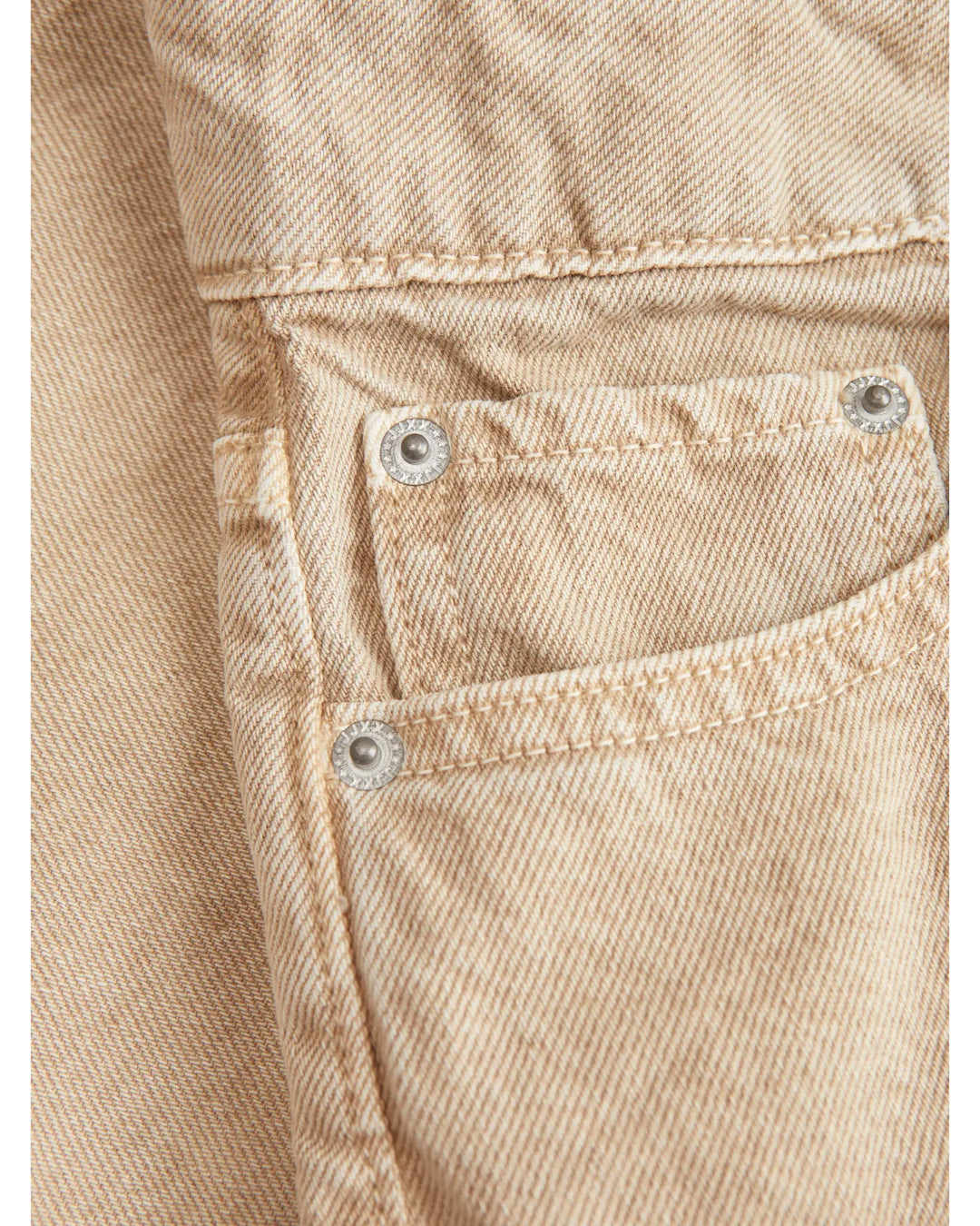 En beige denimlomme med synlige hvide stikninger og tre metalknapper sidder på stoffet. Stoffet er tæt vævet og har en diagonal tekstur, typisk for denim.