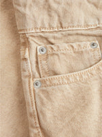 En beige denimlomme med synlige hvide stikninger og tre metalknapper sidder på stoffet. Stoffet er tæt vævet og har en diagonal tekstur, typisk for denim.