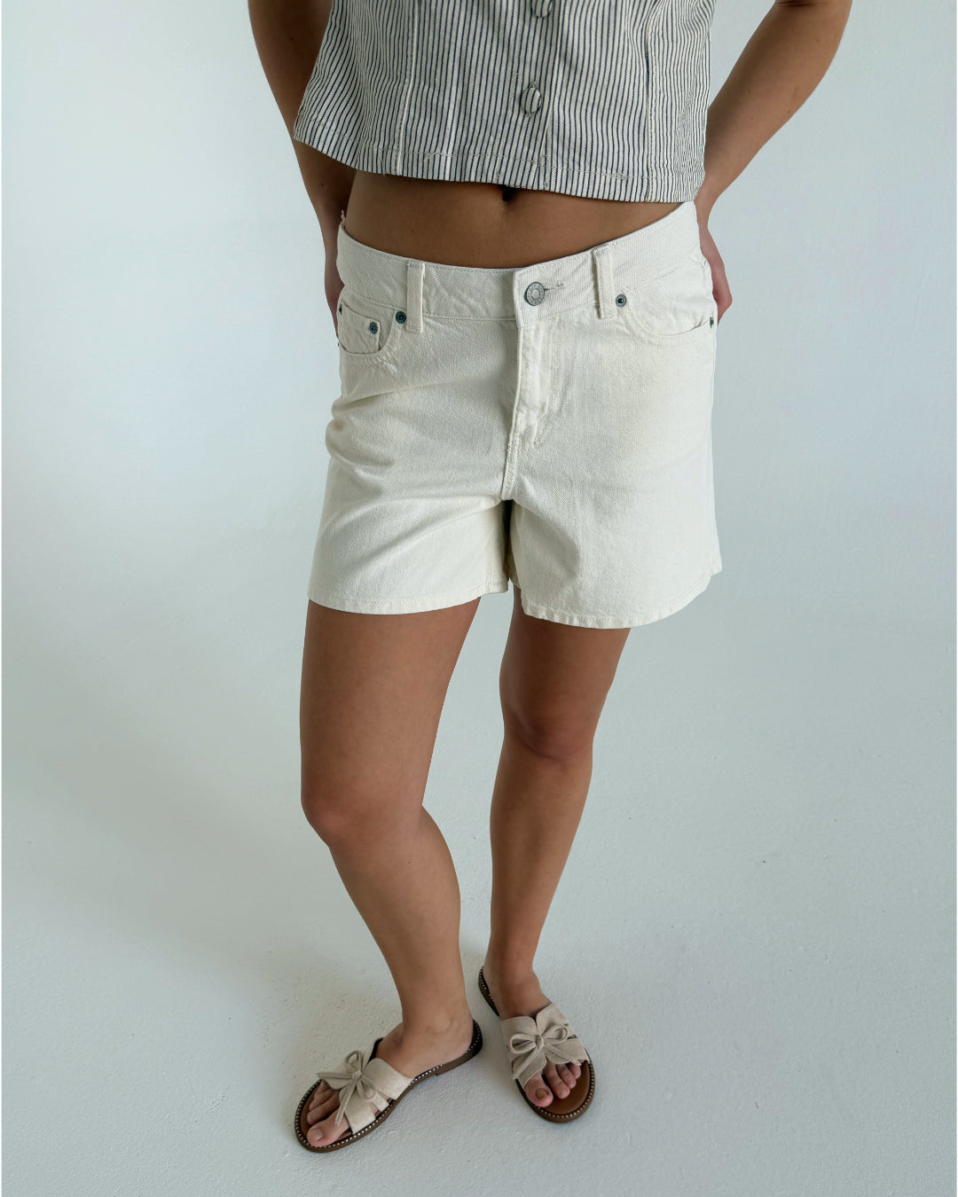 En person bærer hvide shorts og sandaler med sløjfer, mens de står med hænderne i lommerne, iført en stribet skjorte i et lysegråt rum.