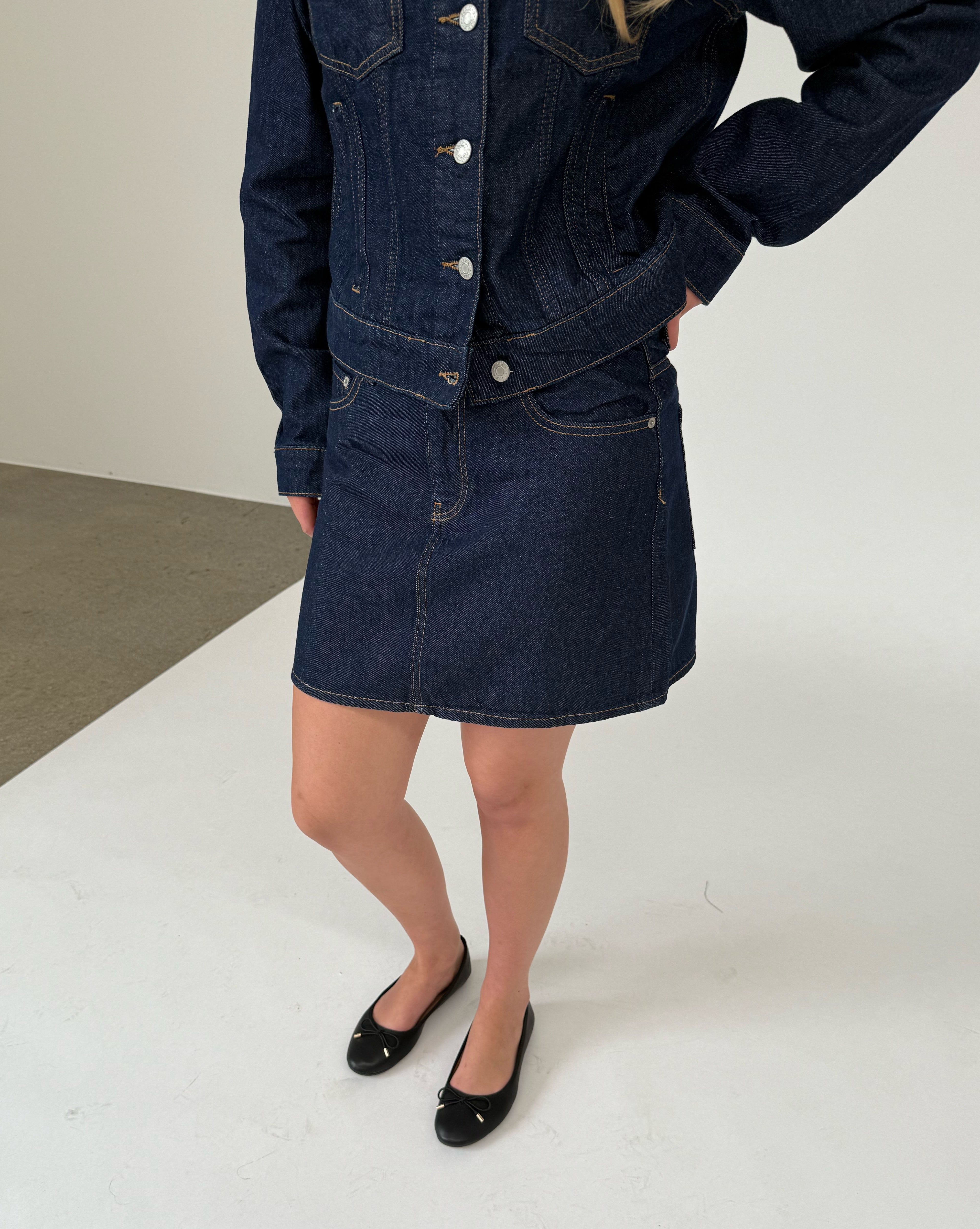 En person i en denimjakke og nederdel står med hænderne i siderne. Personen bærer sorte flade sko. Baggrunden er lys og minimalistisk.