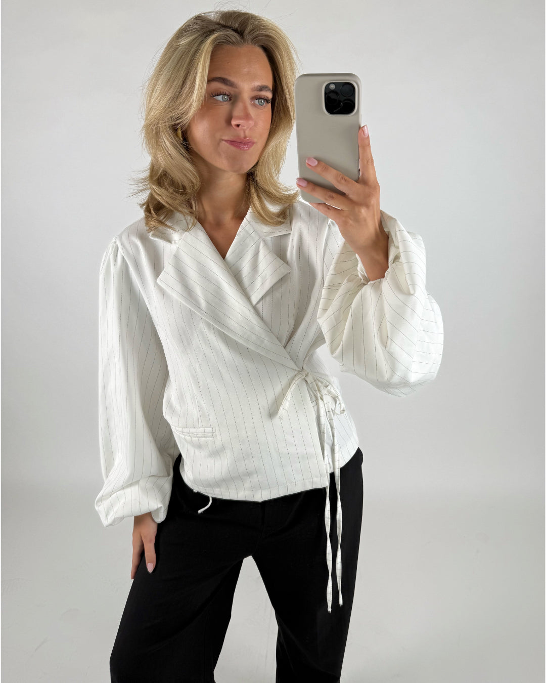 En person iført en hvid slå-om-bluse og sorte bukser tager en selfie med en smartphone. Baggrunden er en simpel, lys væg.