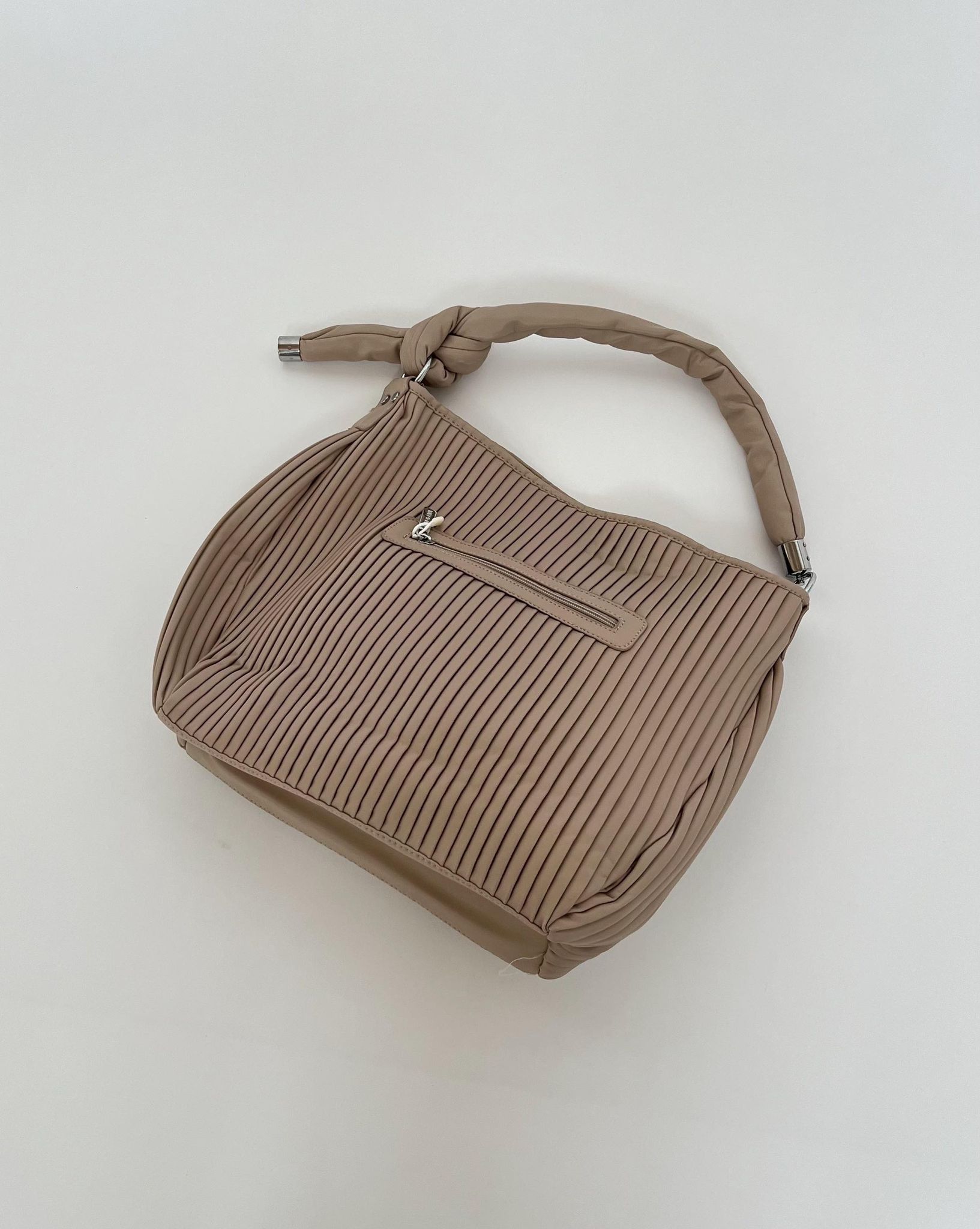 En beige håndtaske med plisseret design, har en lynlås lomme på ydersiden. Håndtaget er knyttet og posen ligger på en hvid baggrund.