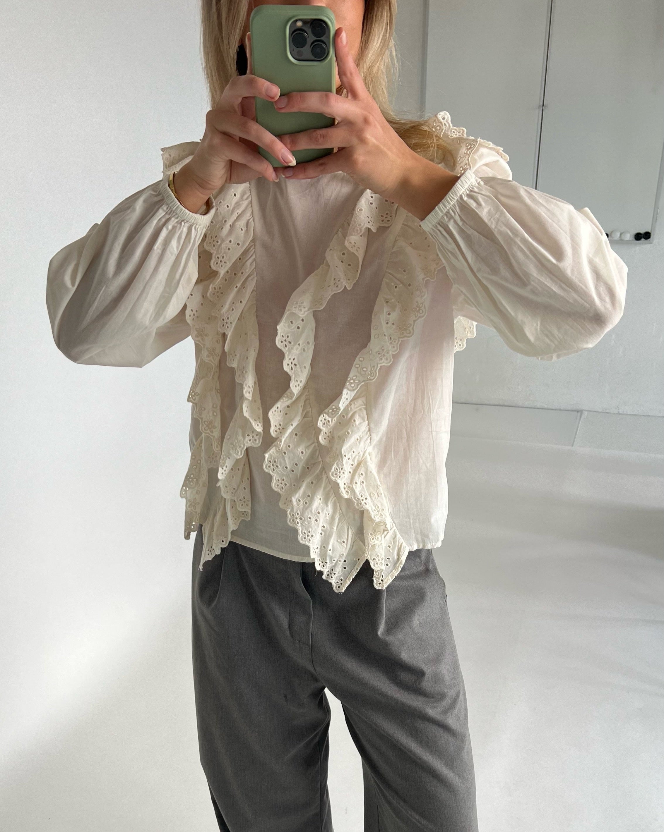 En person tager et spejlfoto iført en hvid bluse med blondekanter og grå bukser, mens de holder en grøn telefon. Baggrunden er minimalistisk og lys.