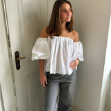 En person står ved en hvid dør iført en hvid off-shoulder bluse og grå bukser. Rummet er lyst med en enkel indretning.