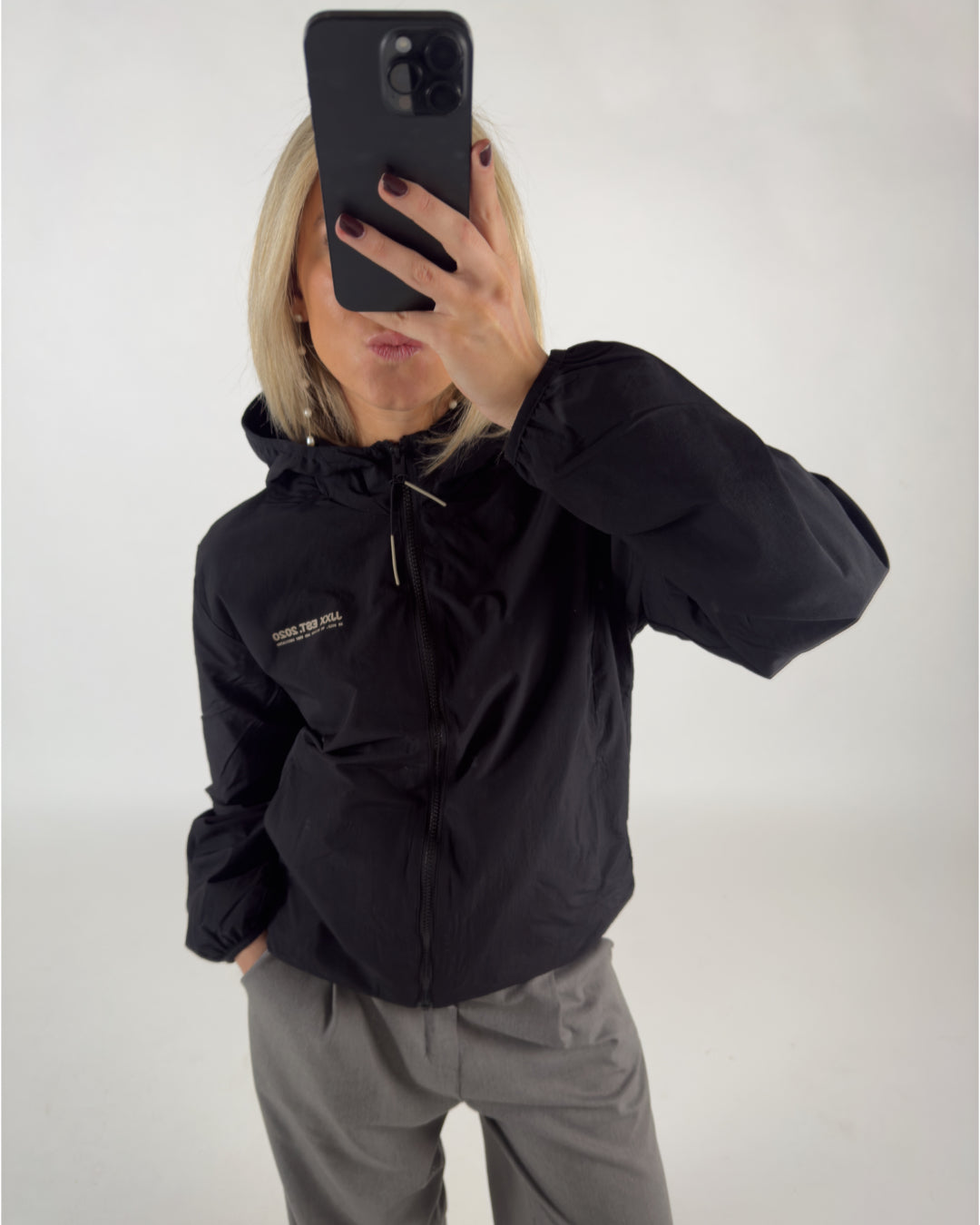 En person tager en selfie med en smartphone og bærer en sort jakke med teksten "GOOSE THE KILL INTERNATIONAL" i hvid. Baggrunden er ensfarvet lys.