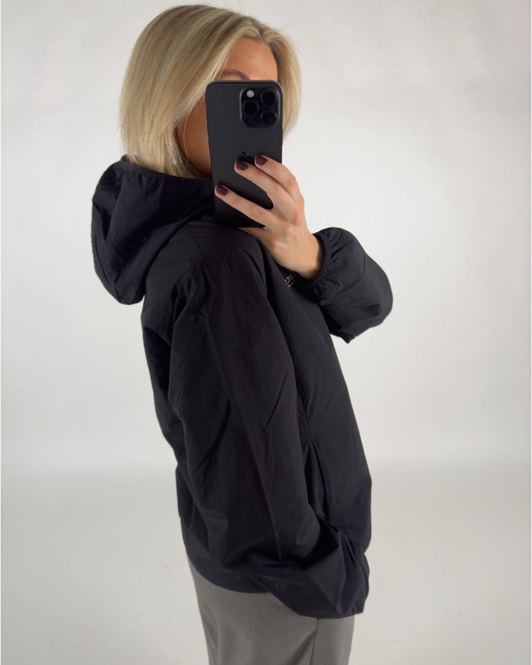 Person iført sort jakke tager selfie med sort telefon, står i neutralt studio.