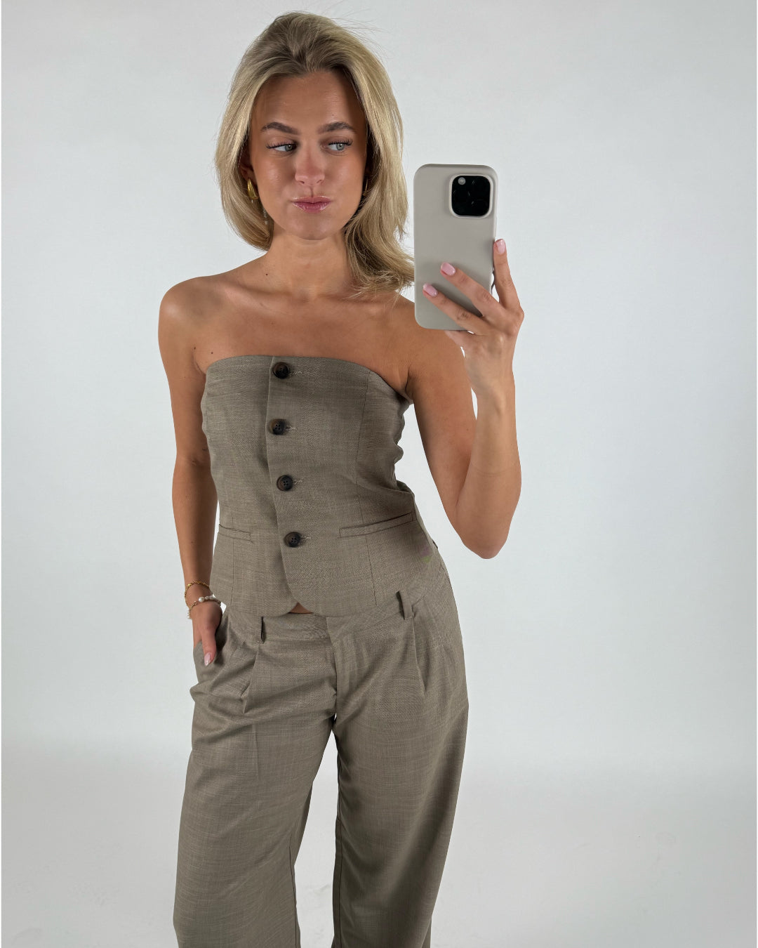 Kvinden tager en selfie iført en beige, ærmeløs top med knapper og bukser i et lyst rum. Hun holder en telefon foran et spejl med en neutral baggrund.