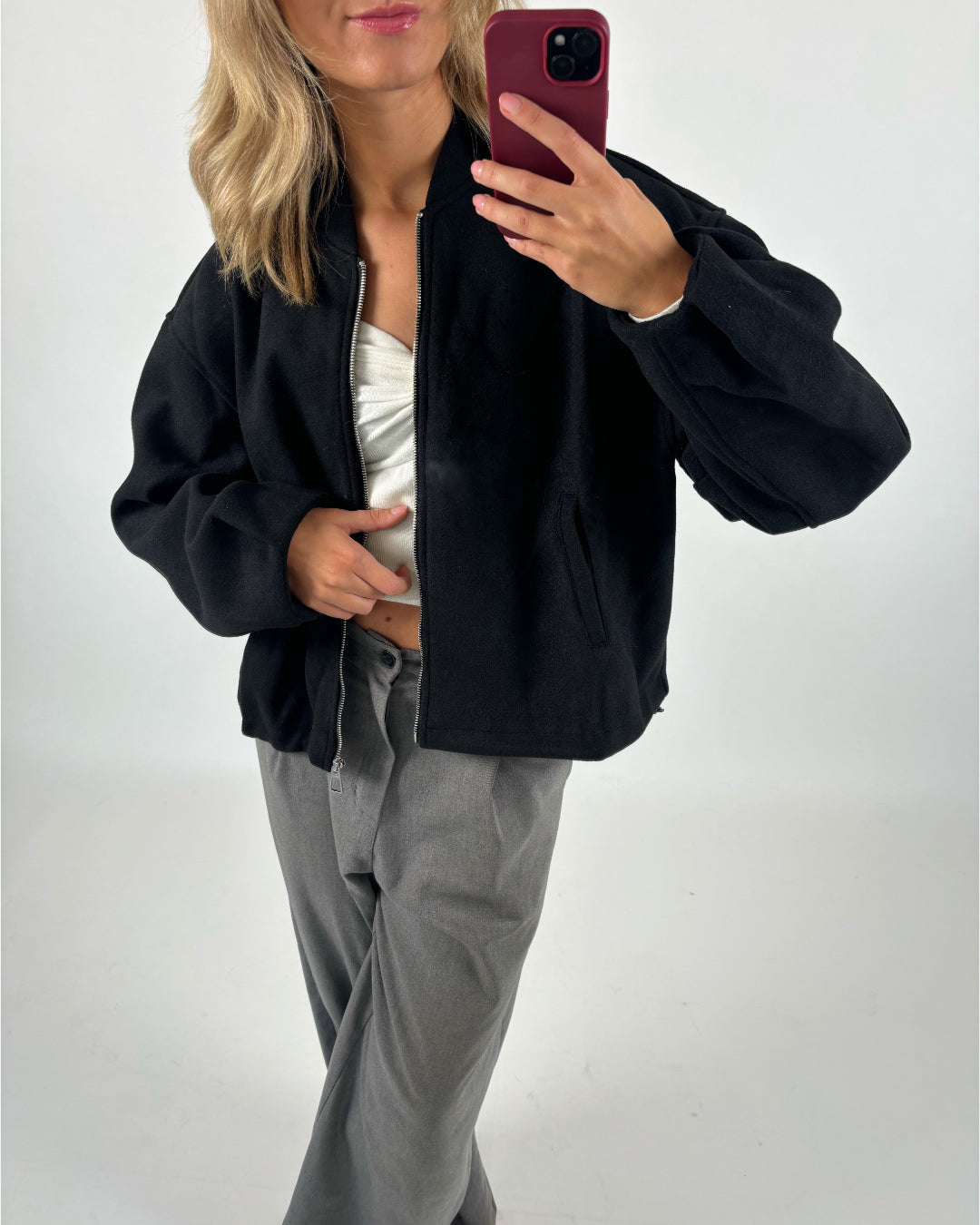 Person tager et selfie iført en sort jakke og grå bukser, stående i et lyst rum. Jakken er åben, afslørende en hvid top. Personen holder en telefon med rød etui.