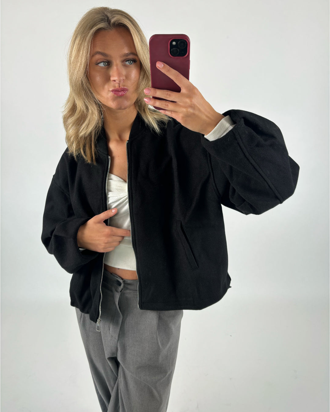En person tager en selfie med en smartphone iført en sort jakke og grå bukser. Personen står foran en lys, ensfarvet baggrund.