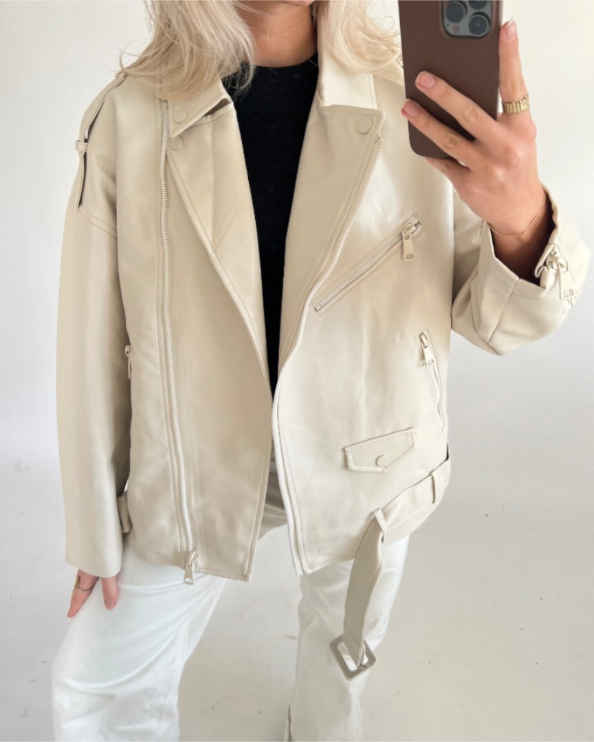 En person iført en beige læderjakke og hvide bukser tager et spejlbillede med en smartphone i en minimalistisk, lys baggrund.