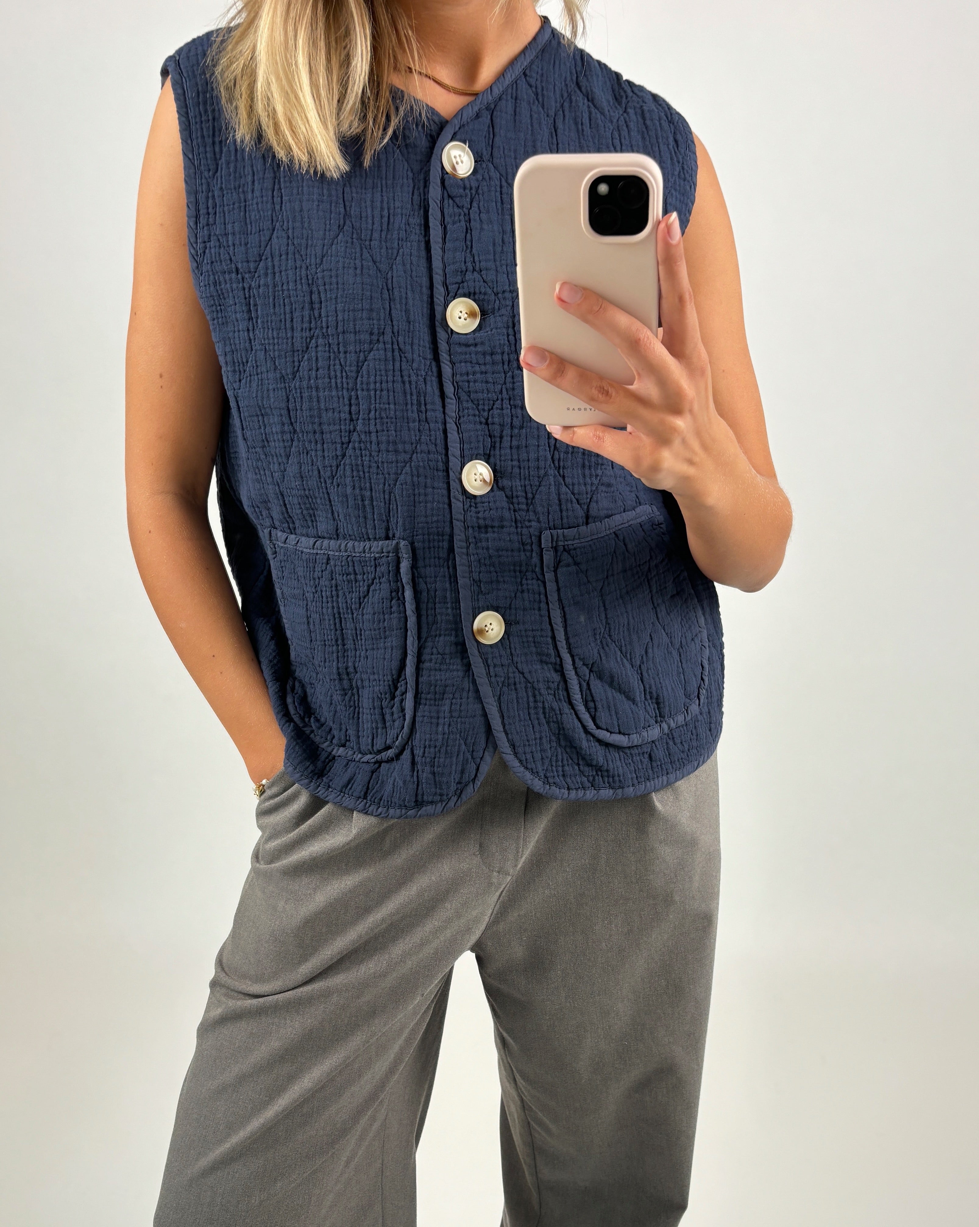 En person iført en mørkeblå quiltet vest fotograferer sig selv med en smartphone. Vesten har store knapper og lommer, og personen står i grå bukser mod en lys baggrund.