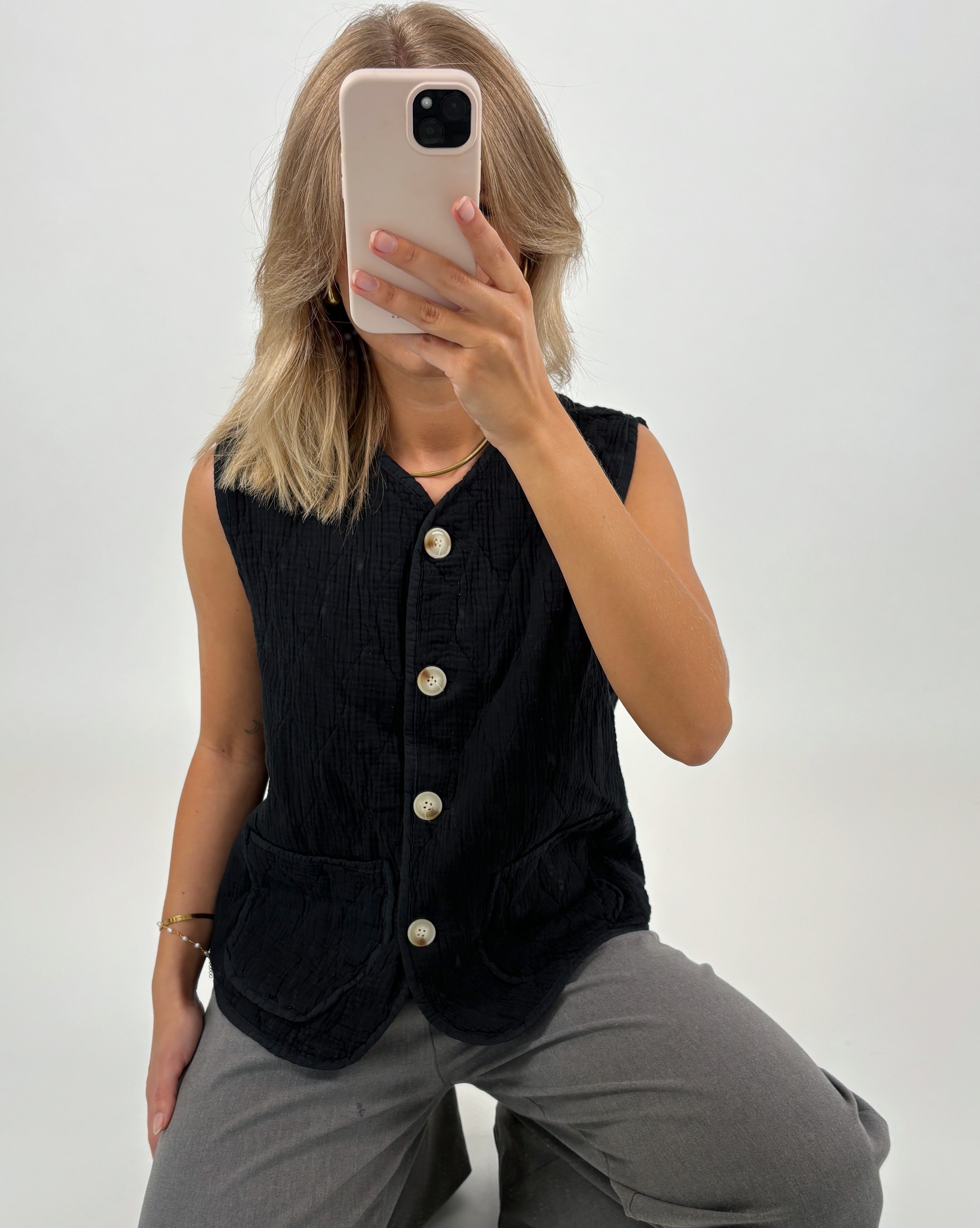 En person tager et selfie, iført en sort vest med knapper og grå bukser, mod en lys baggrund.
