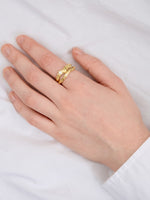 Hånd bærer en gylden ring med tekstur på fjerde finger, mens hånden hviler på en hvid overflade, skaber en elegant og enkel scene.