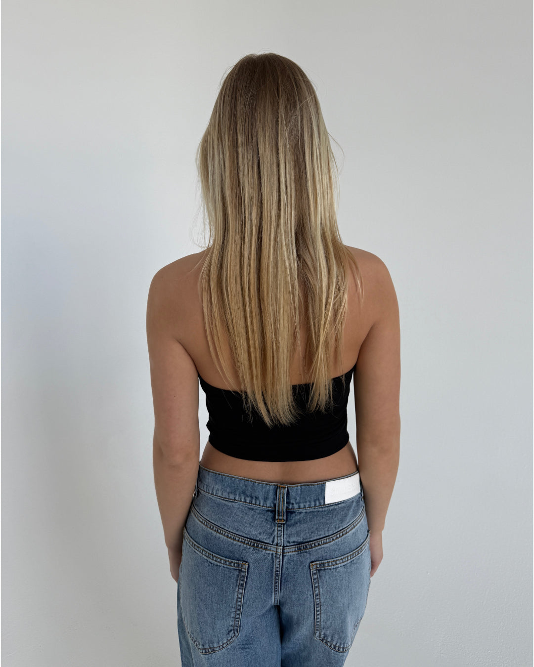 Kvinden har langt, lyst hår og bærer en sort, stropløs top samt blå jeans. Hun står med ryggen til i et rum med en ensfarvet, hvid væg.