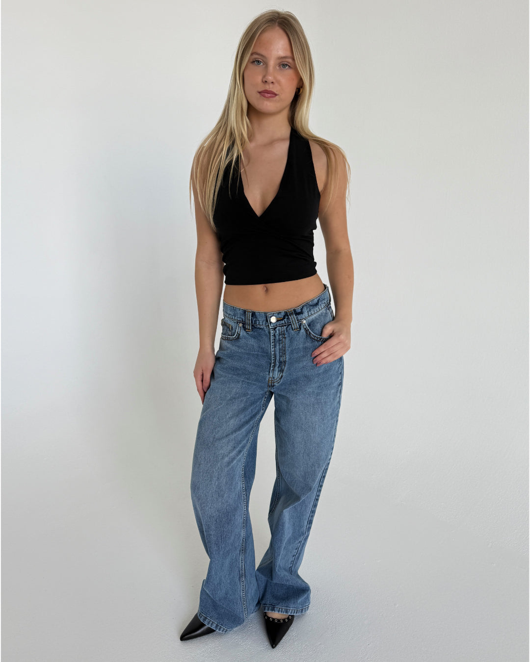 En person står afslappet iført en sort top og blå jeans i et lyst rum. Personen hviler venstre hånd i jeanslommen og har på sig sorte, spidse sko.