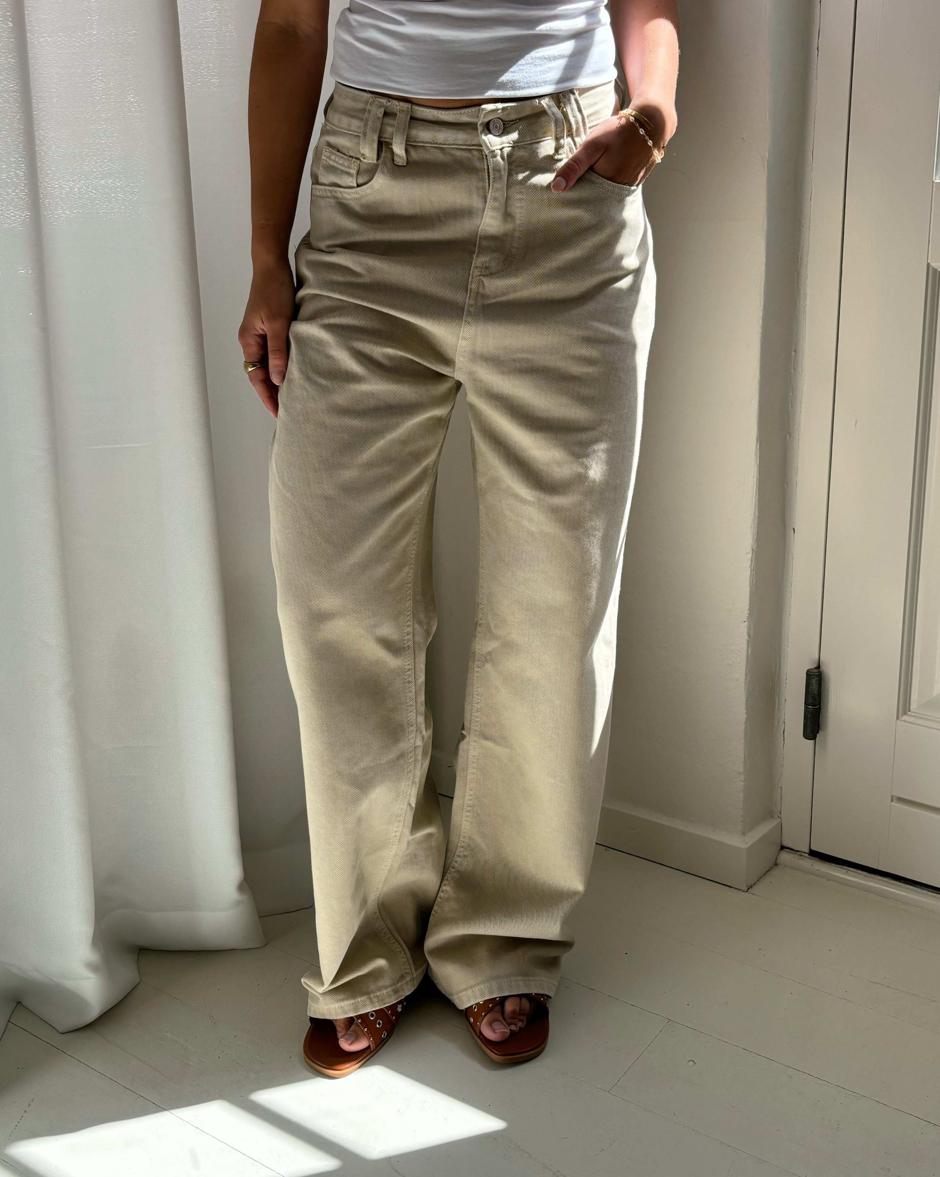 Person iført beige bukser og hvid tanktop står afslappet med hænderne i lommerne, i et rum med lys baggrund, gardiner og dør.
