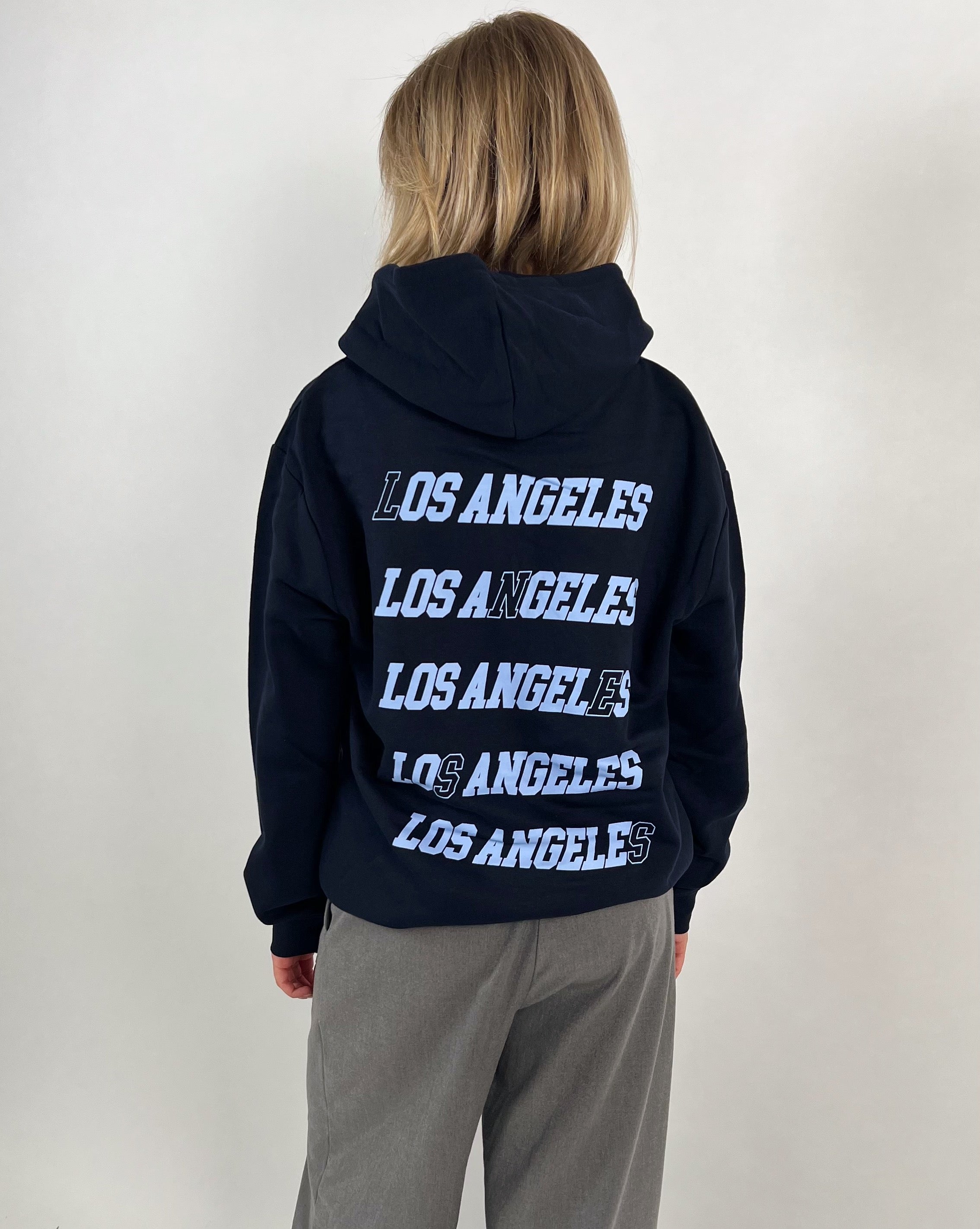 En person står med ryggen til iført en sort hættetrøje med teksten "LOS ANGELES" gentaget fem gange i hvid tekst. Baggrunden er lys og ensfarvet.