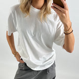 Person iført hvid t-shirt og grå bukser tager et selfie med brun mobiltelefon foran en neutral baggrund. Hånden i bukselommen, anden hånd holder telefonen.