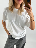 Person iført hvid t-shirt og grå bukser tager et selfie med brun mobiltelefon foran en neutral baggrund. Hånden i bukselommen, anden hånd holder telefonen.