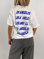 En person bærer en hvid T-shirt, hvorpå der står "LOS ANGELES" gentaget fire gange i blå skrift. Personen står med ryggen til i et neutralt rum.