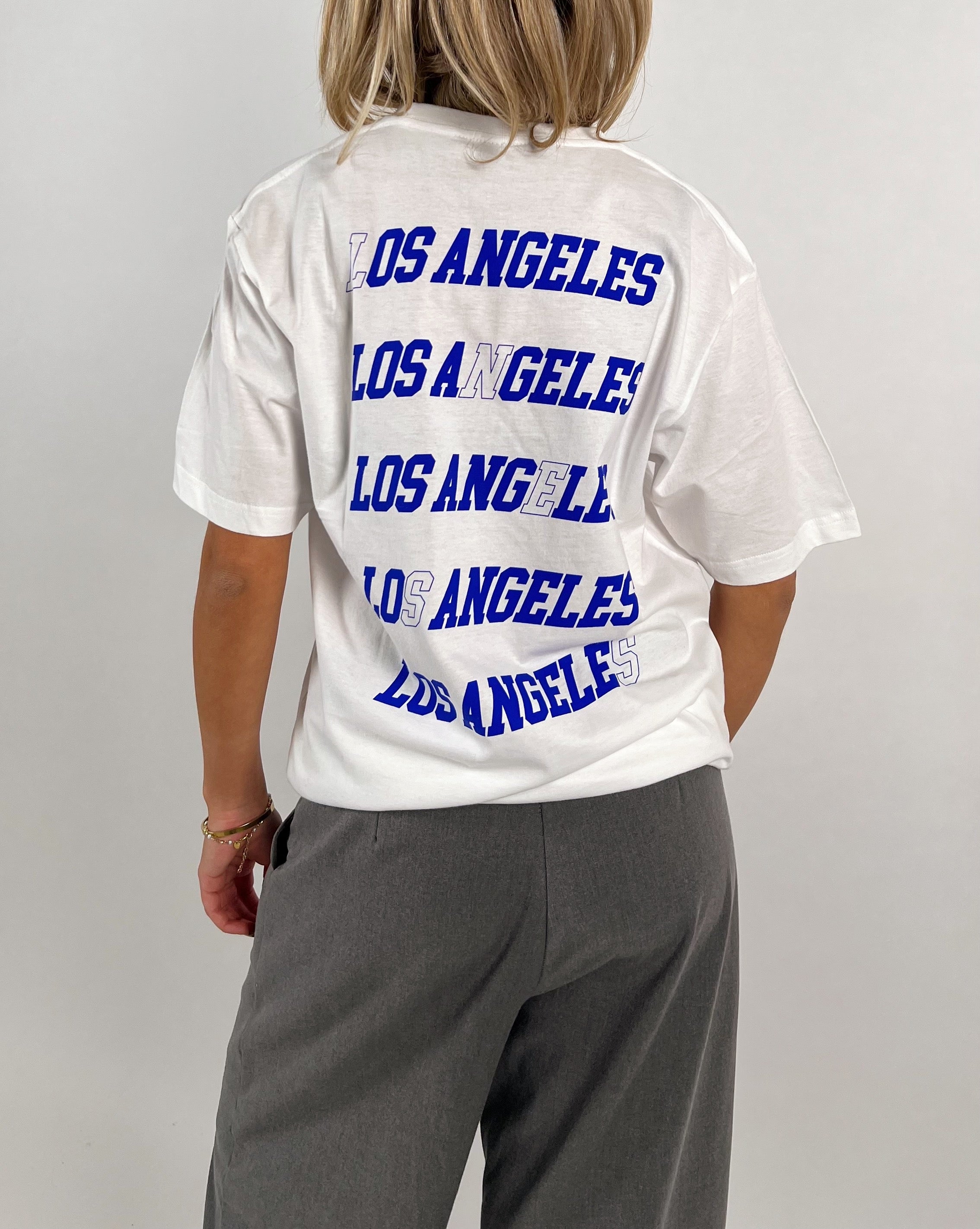 En person bærer en hvid T-shirt, hvorpå der står "LOS ANGELES" gentaget fire gange i blå skrift. Personen står med ryggen til i et neutralt rum.