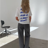 En person med langt hår iført en hvid T-shirt med teksten "LOS ANGELES" flere gange på ryggen, står i et lyst rum med en sort stol ved siden af.