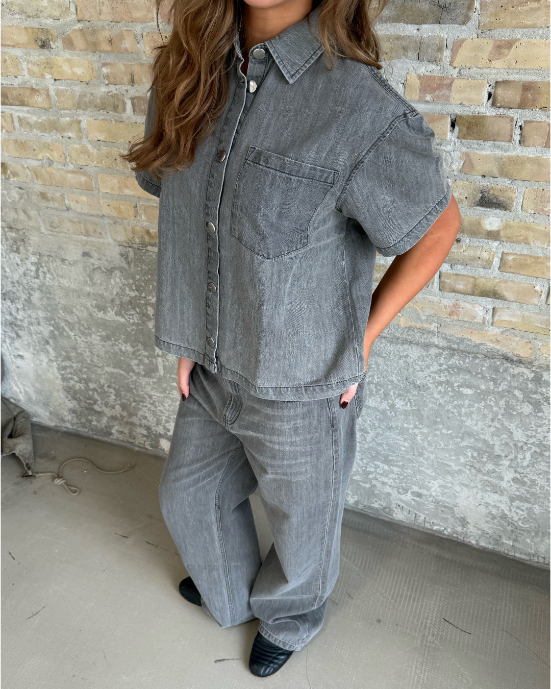 En person iført gråt denim outfit står afslappet mod en rå murstensvæg. Personens hænder er i lommerne, og de bærer sorte sko på et gråt betongulv.