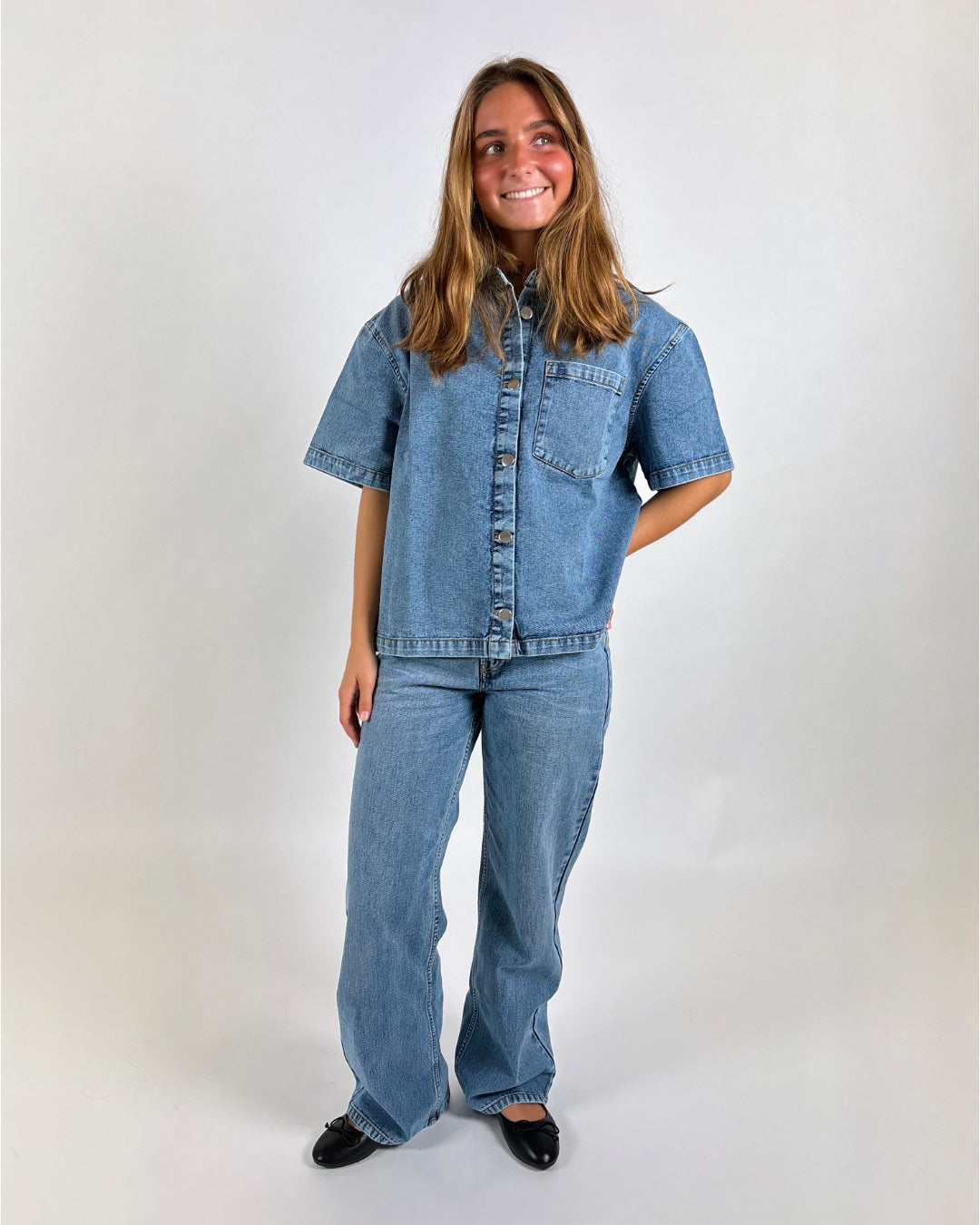 En person står iført en denimskjorte og jeans, hænderne i lommerne, smilende. Baggrunden er en enkel, lys væg, der fremhæver påklædningen.