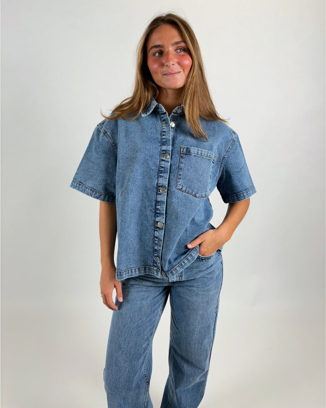 En person står iført en denimskjorte og jeans, kigger lidt til siden, med en lille smil. Baggrunden er simpel og lys, hvilket fokuserer på tøjet.