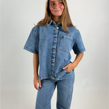 En person står iført en denimskjorte og jeans, kigger lidt til siden, med en lille smil. Baggrunden er simpel og lys, hvilket fokuserer på tøjet.