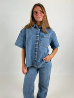 En person står iført en denimskjorte og jeans, kigger lidt til siden, med en lille smil. Baggrunden er simpel og lys, hvilket fokuserer på tøjet.