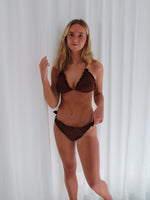 En person iført brun bikini står stille foran et hvidt gardin og kigger mod kameraet. Stemningen i rummet er lys og minimalistisk.
