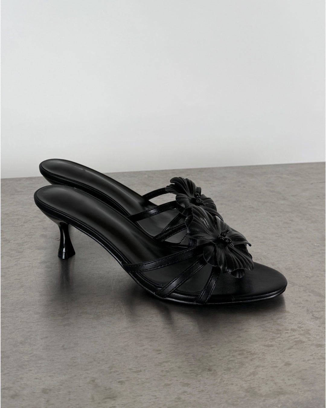 Sort sandal med høje hæle og blomsterdetaljer står på en grå overflade.