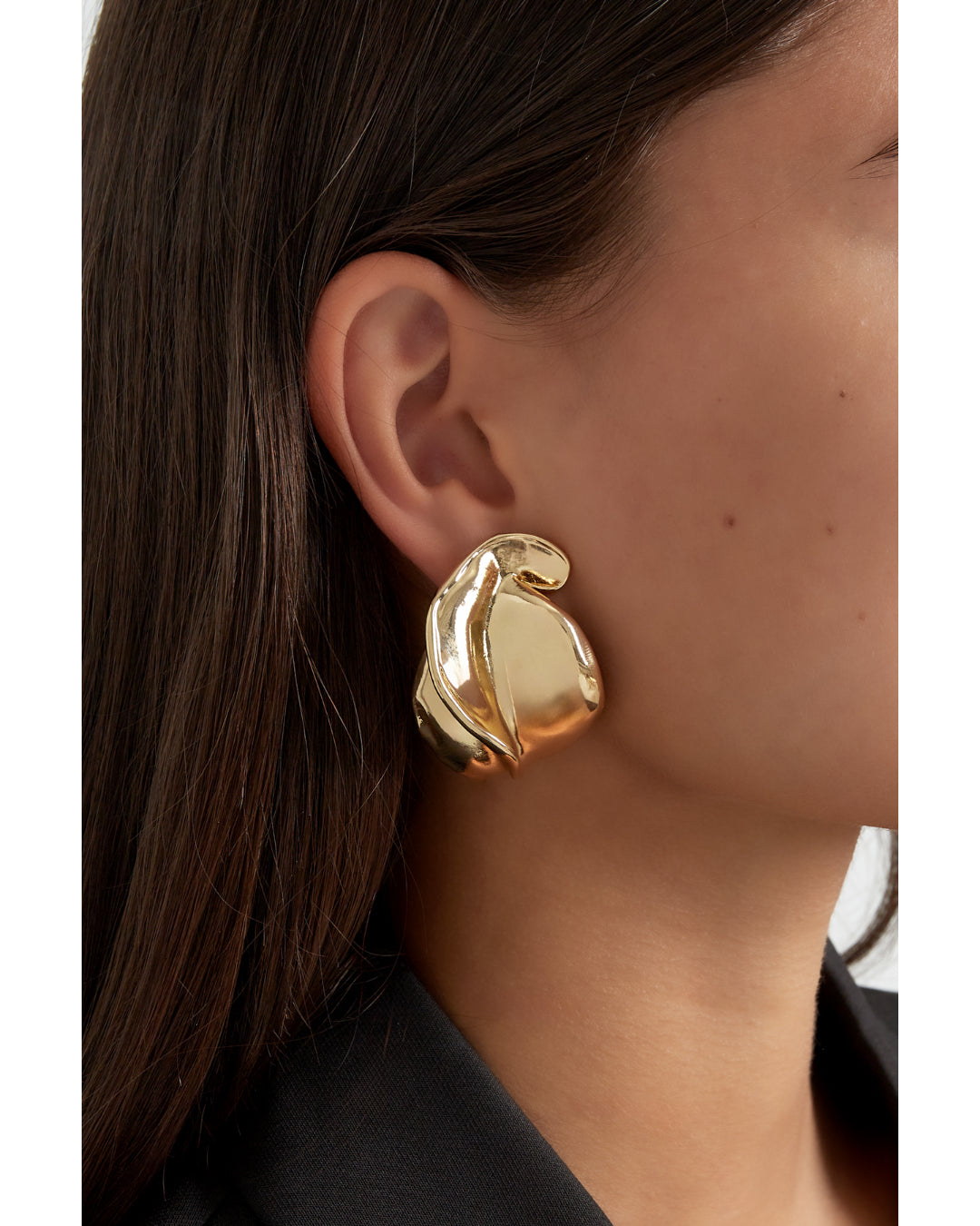 Ørering, skinnende og bølgende, hænger fra øret. Personen har mørkt hår og bærer en sort top, hvilket fremhæver smykkets gyldne glans i en elegant stil.