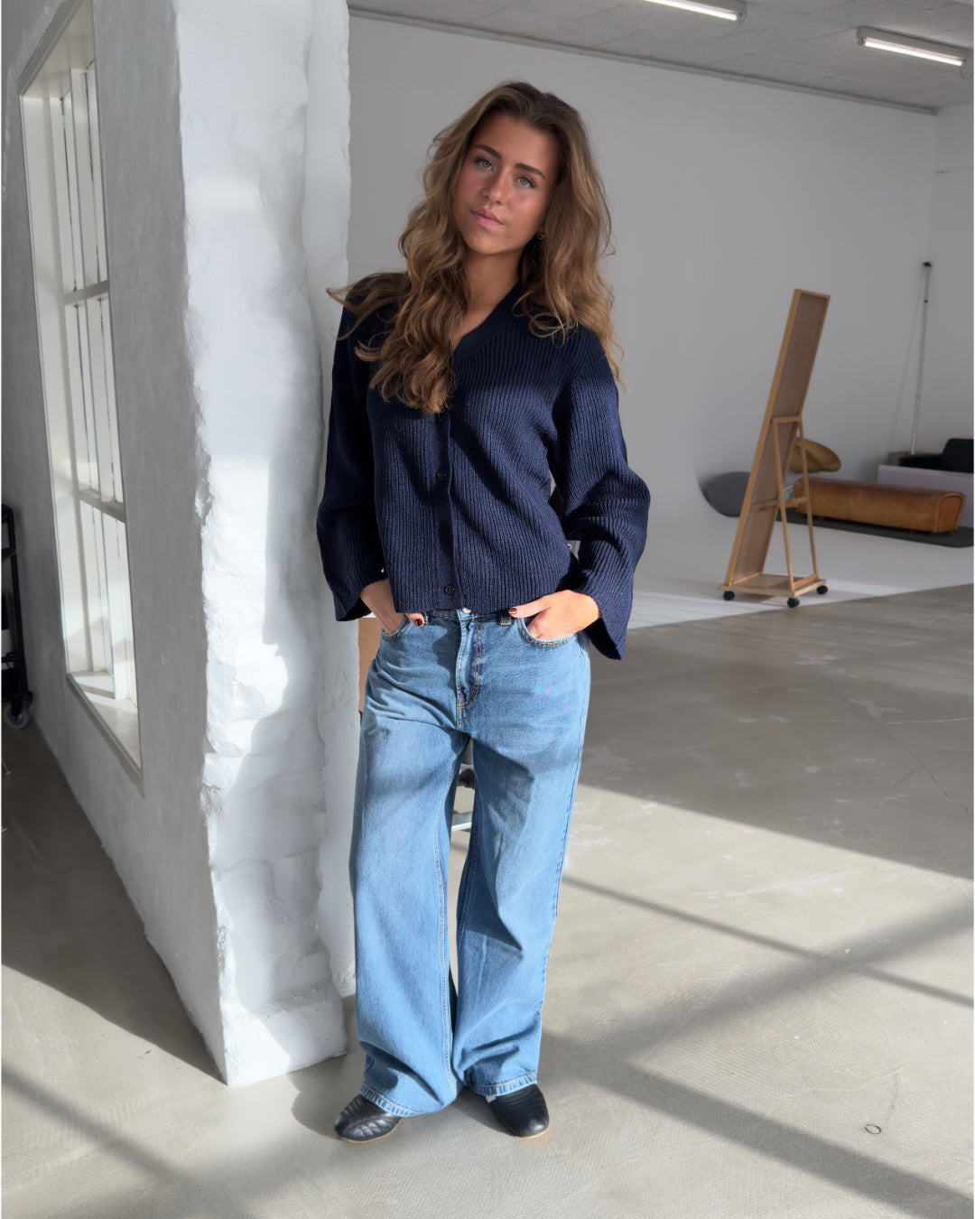 Kvinden står afslappet op ad en hvid mur, iført en mørk trøje og lyse jeans. Rummet har store vinduer og moderne indretning med minimalisme som stil.