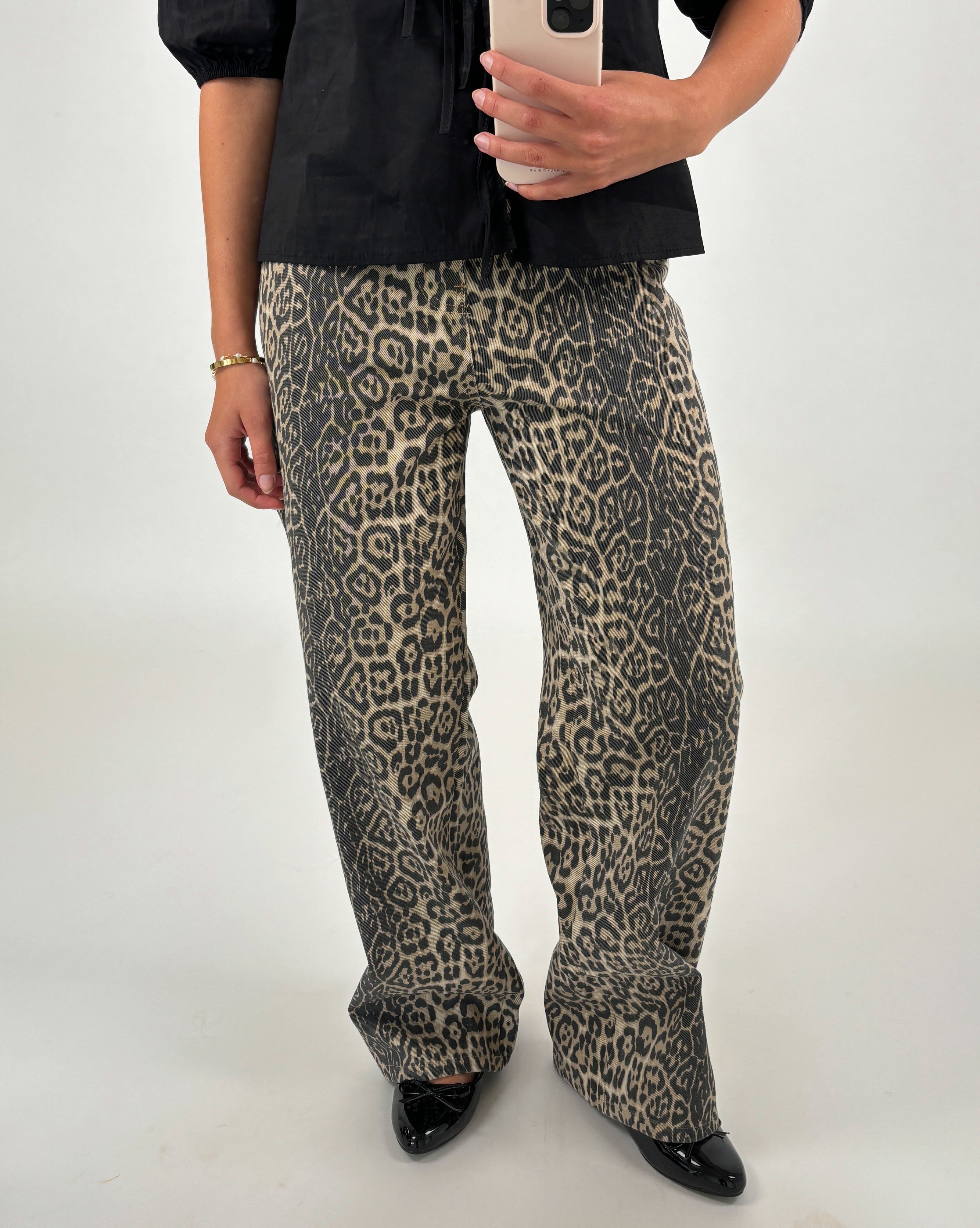 En person fotograferer sig selv iført leopardmønstrede bukser og en sort bluse, mens de står i et lyst rum. Personen holder en smartphone i den ene hånd.