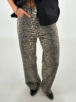 Bukser med leopardprint bæres af en person, der hviler en hånd i lommen. Personen har sorte sko og en sort overdel. Baggrunden er neutral og lys.
