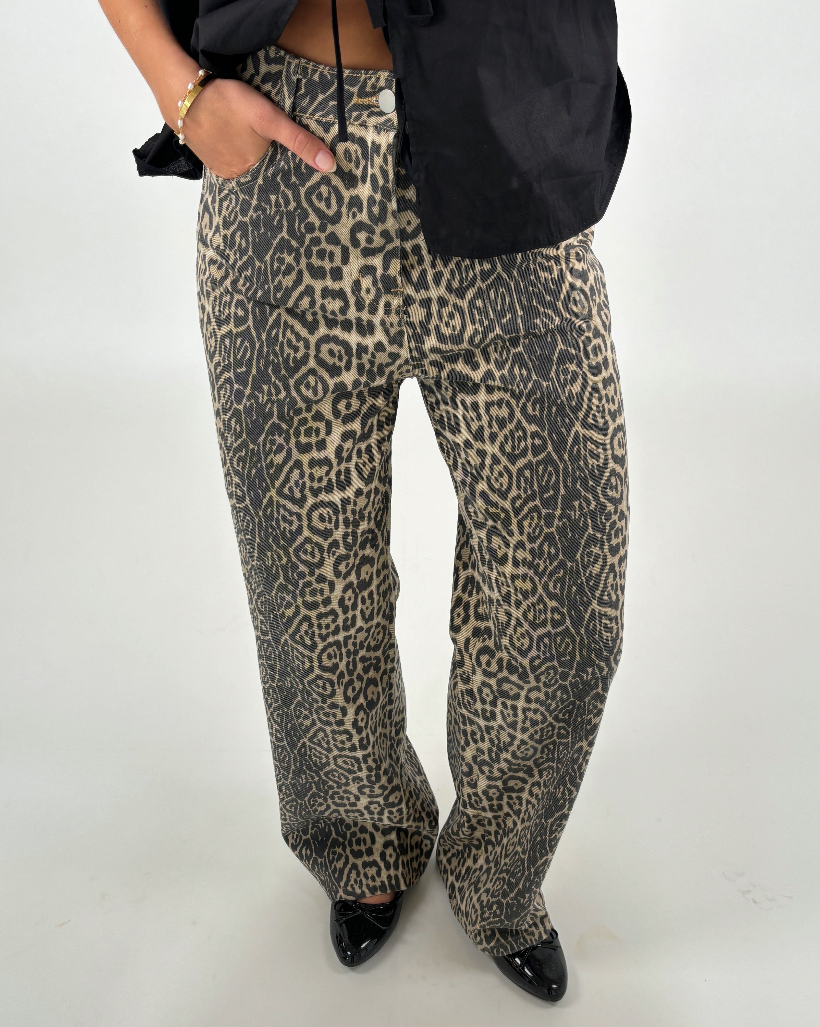 Bukser med leopardprint bæres af en person, der hviler en hånd i lommen. Personen har sorte sko og en sort overdel. Baggrunden er neutral og lys.