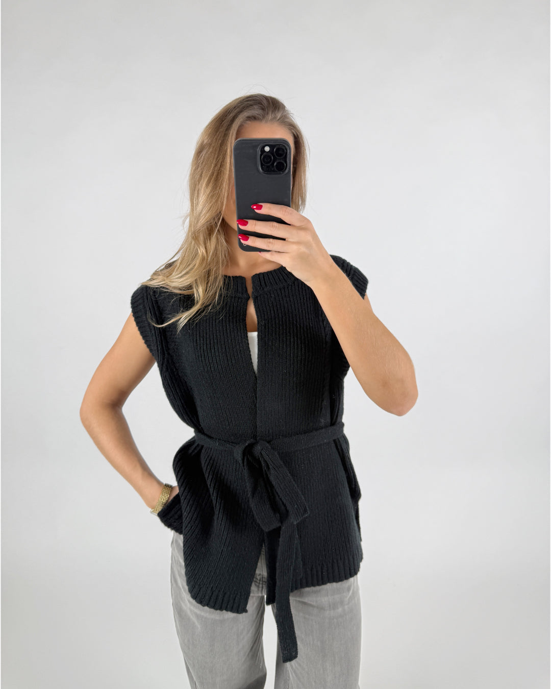 Person i sort vest tager selfie med mobiltelefon mod en hvid baggrund. De bærer grå bukser og har hånden på hoften, viser rød neglelak.