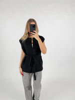 En person iført sort ærmeløs sweater og grå bukser tager et billede med en mobiltelefon i en simpel, hvid baggrund.