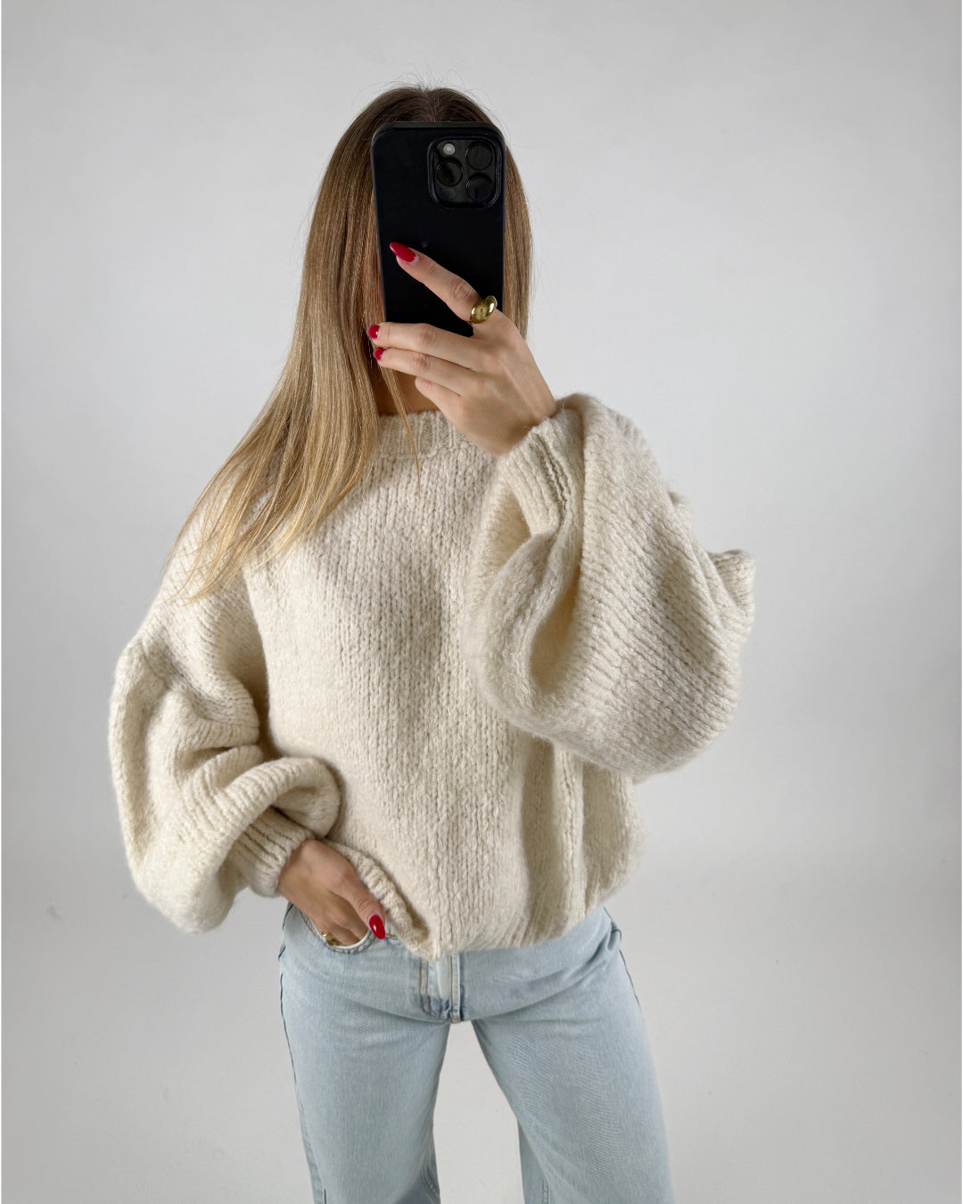 En person iført en cremefarvet, voluminøs sweater holder en mobiltelefon op til ansigtet. Vedkommende har lange lysebrune hår og står i et simpelt rum med lyseblå jeans.