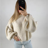En person iført en cremefarvet, voluminøs sweater holder en mobiltelefon op til ansigtet. Vedkommende har lange lysebrune hår og står i et simpelt rum med lyseblå jeans.