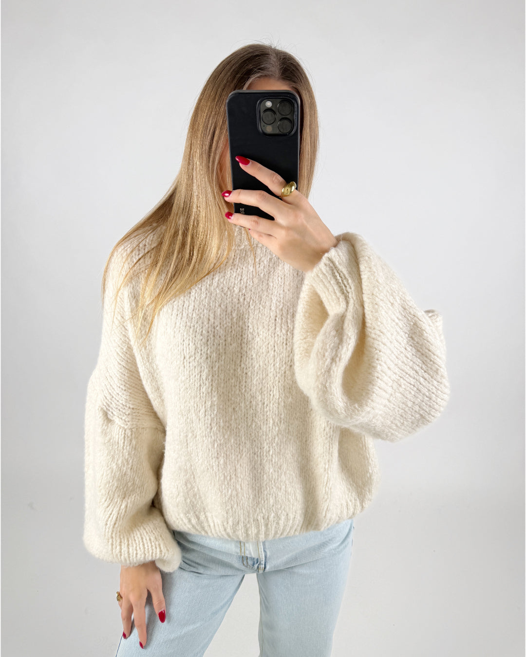 En person iført en hvid, strikket sweater holder en mobiltelefon med sort cover foran ansigtet; personen har lange, lyse lokker, og neglene er rødmalede. Baggrund er ensfarvet lys.