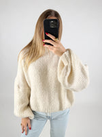 En person iført en hvid, strikket sweater holder en mobiltelefon med sort cover foran ansigtet; personen har lange, lyse lokker, og neglene er rødmalede. Baggrund er ensfarvet lys.
