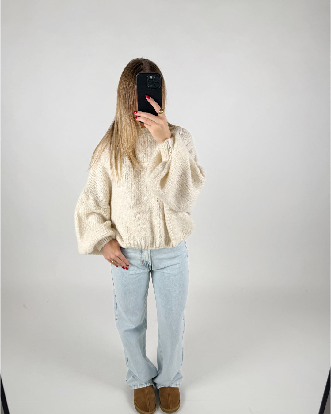 En person står i et lyst rum, tager et spejlbillede. De bærer en stor, hvid sweater, lyse jeans og brune sko, mens de holder en mørk telefon foran ansigtet.