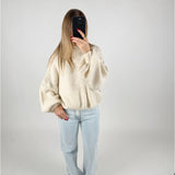 En person står i et lyst rum, tager et spejlbillede. De bærer en stor, hvid sweater, lyse jeans og brune sko, mens de holder en mørk telefon foran ansigtet.