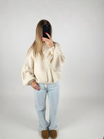 En person står i et lyst rum, tager et spejlbillede. De bærer en stor, hvid sweater, lyse jeans og brune sko, mens de holder en mørk telefon foran ansigtet.