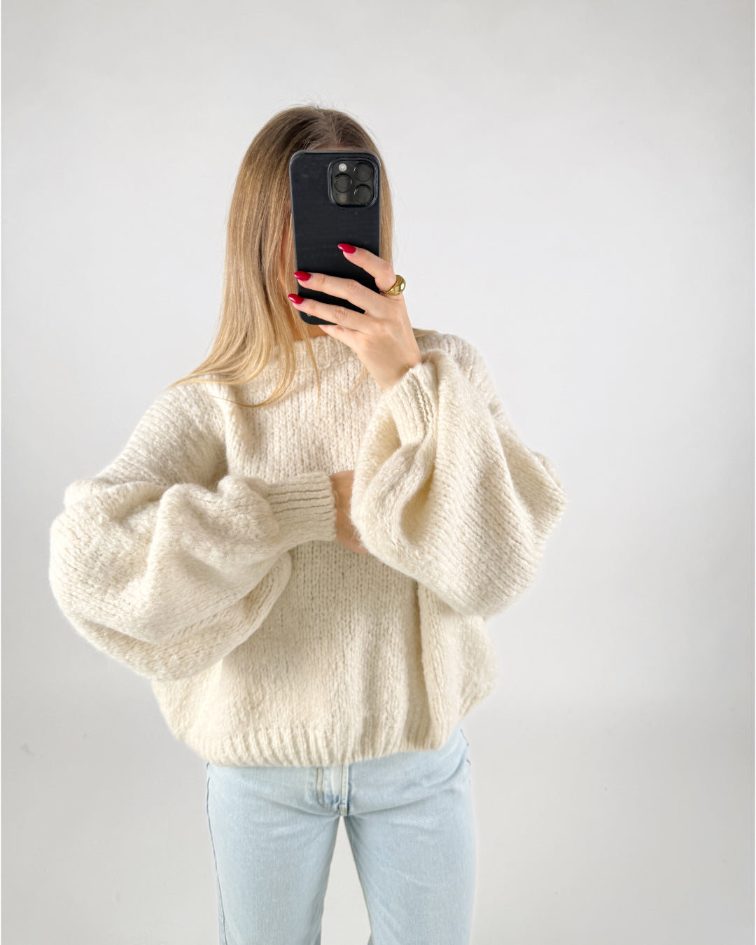 Person tager et billede med en smartphone, iført en stor, lys sweater. Personen har løst, langt hår og røde negle. Baggrunden er en simpel, hvid væg.