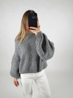 Kvinde iført grå striktrøje tager et spejlbillede med en sort telefon. Hun står i et minimalistisk rum med lys baggrund, iført hvide bukser.