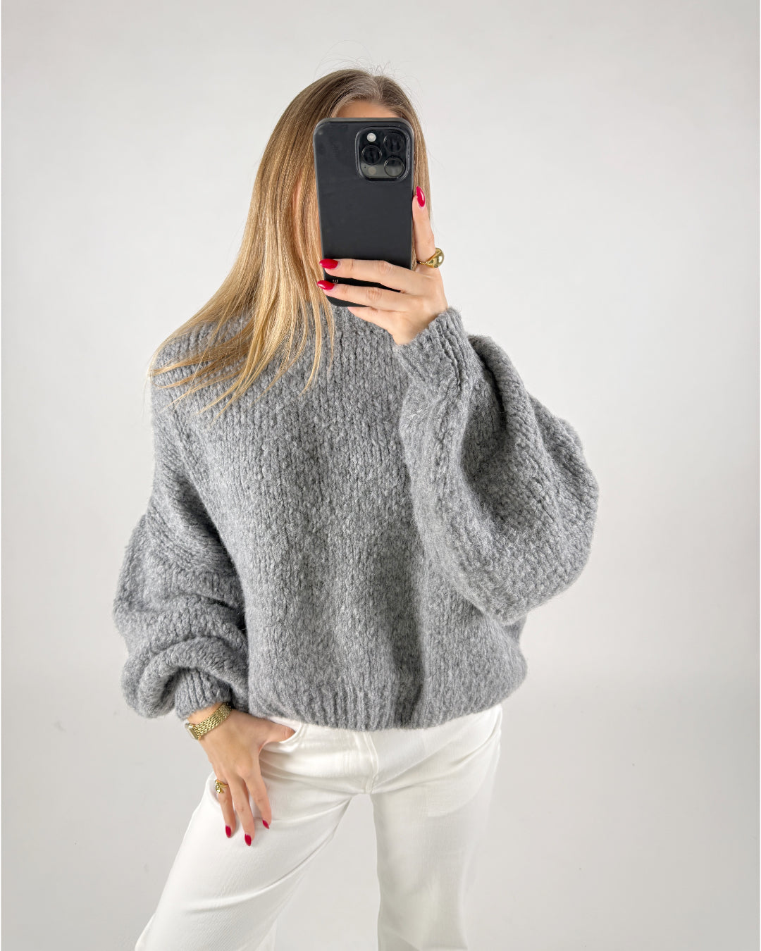 En person iført en grå sweater tager en selfie med en sort smartphone. Personen har hvidt tøj på og rød neglelak. Baggrunden er ensfarvet hvid.